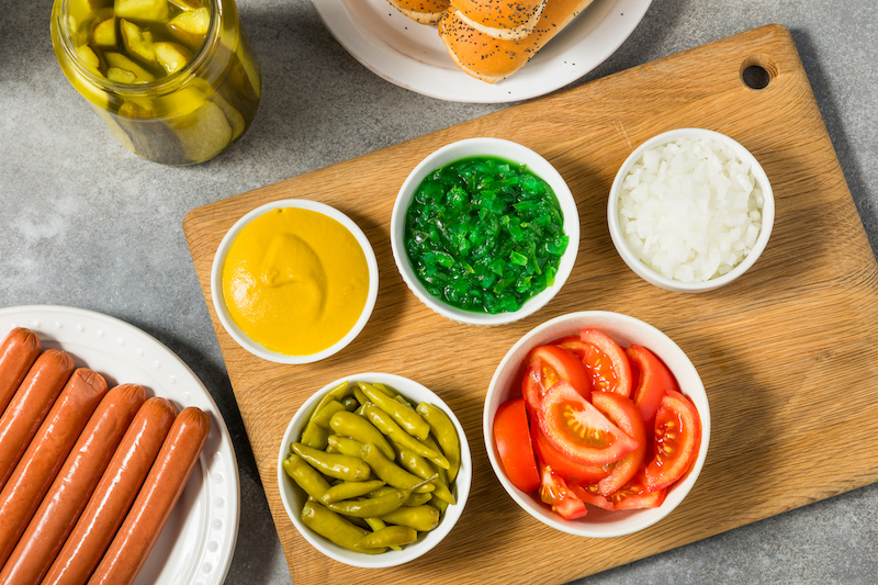 Hot dog toppings including mustard, relish, onions, tomatoes, and peppers on a cutting board