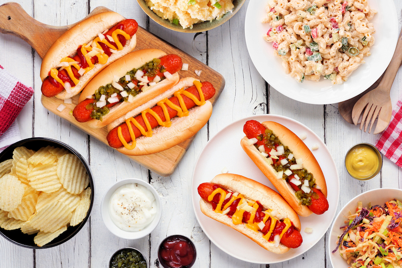 Plates of hot dogs with mustard and relish surrounded by salads, chips, and condiments
