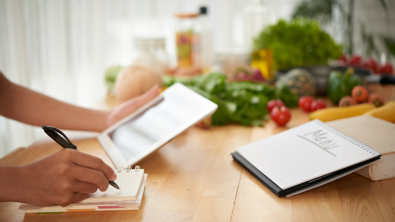 Person planning meals using a tablet and notebook on a kitchen counter with fresh ingredients