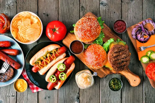 Burgers and hot dogs with buns, chips, and assorted toppings on a wooden table