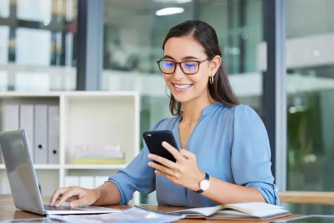 Woman using her phone while working on a laptop at a desk