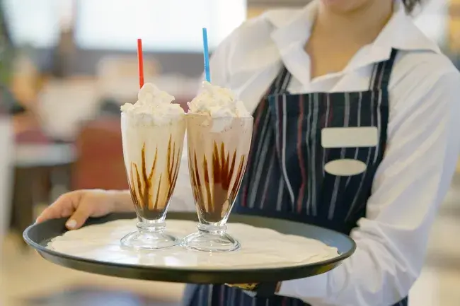 Two ice cream malts topped with whipped cream being served by a waiter on a tray