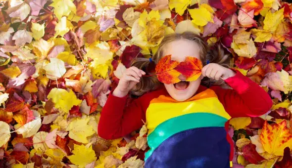 Child lying in autumn leaves holding red leaves over their eyes