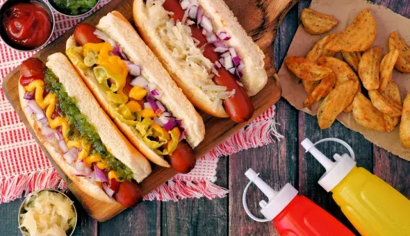 Burgers and hot dogs with buns, chips, and assorted toppings on a wooden table