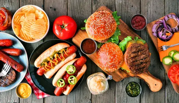 Burgers and hot dogs with buns, chips, and assorted toppings on a wooden table