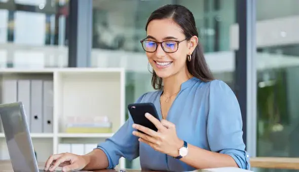 Woman using her phone while working on a laptop at a desk
