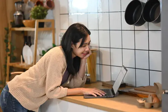 Smiling woman using laptop on kitchen counter with cookware in background